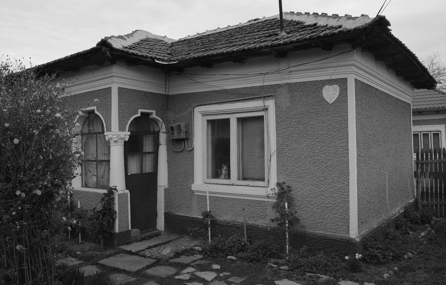 Detaliu cu fațada tencuită a unei locuințe, cu decorații în relief; intrarea se face printr-o verandă cu un stâlp cu capitel decorat. La fereastră este așezată o pisică privind spre obiectiv..